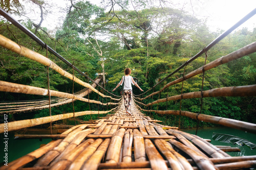 Back view of young woman on suspension wooden bamboo bridge across Loboc river in jungle. Vacation on tropical island. Bohol, Philippines photo