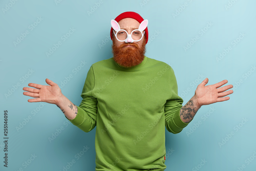 Puzzled guy faces dilemma, spreads hands in uncertainty, prepares for carnival, has image of hare, wears spectacles with rabbit ears, has red thick beard, dressed in green jumper, isolated on blue Puzzled guy faces dilemma, spreads hands in uncertainty, prepares for carnival, has image of hare, wears spectacles with rabbit ears, has red thick beard, dressed in green jumper, isolated on blue