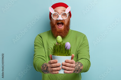 Joyful hipster wears bunny glasses, holds vase with paschal eggs as symbol of new life and resurrection, has fun coloring traitional food for Easter, meets spring holiday. Symbols and traditions photo