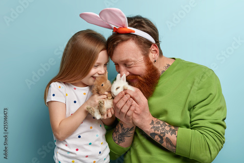 Funny bearded ginger man tells story about Easter bunny to daughter, play with two fluffy decorative rabbits, have happy expressions, rejoice having new pets at home. Family and animals concept