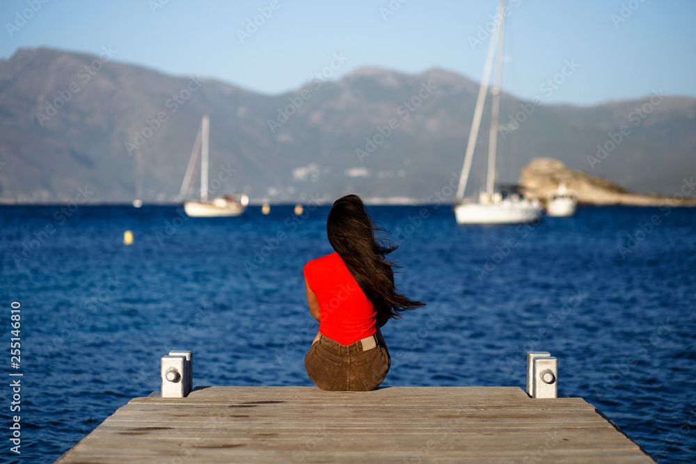A lonely young brunette woman in red sitting with back on wooden pier, admiring seascape of the Corsica island, France. Mountains background. Horizontal view.