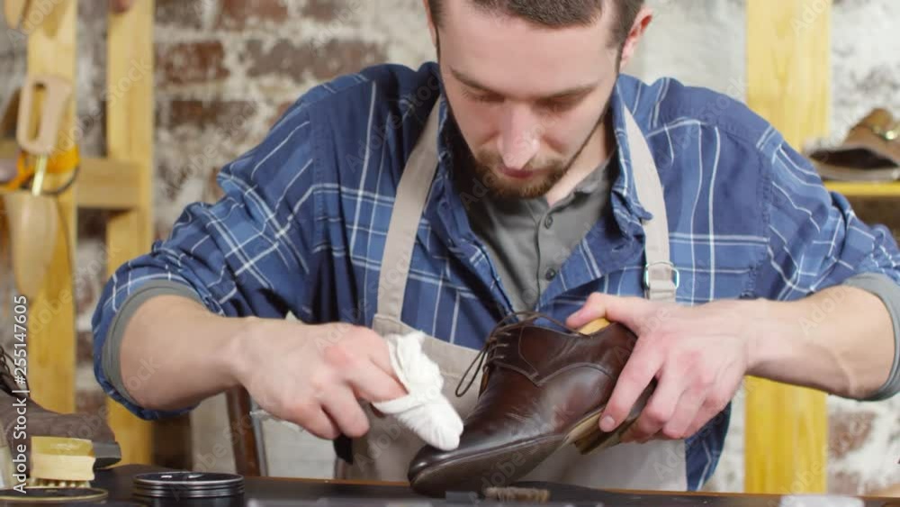 Male shoemaker in apron sitting at desk in workshop and polishing leather boot with cloth wrapped around fingers Male shoemaker in apron sitting at desk in workshop and polishing leather boot with cloth wrapped around fingers