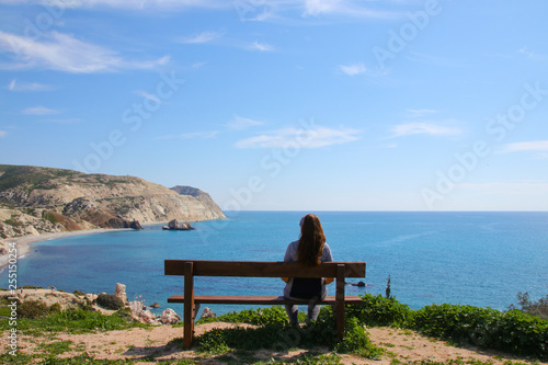 A woman is sitting on a wooden bench and looks at Petra-tou-Romiou - birthplace of Aphrodite on Cyprus, view from nature trail Petra-tou-Romiou