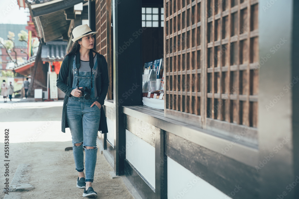 full length young asian woman traveler with backpack and hat looking inside shitennoji temple osaka japan. curious female walking in shinto shrine sightseeing in traditional wooden japanese house.