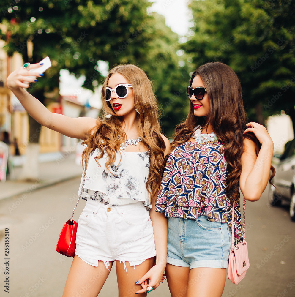 Hipster girls friends standing on road together and taking self portrait on  smart phone. Brunette and blonde having fun, smiling and posing on street.  Wearing sunglasses, jeans shorts and tops. Stock Photo