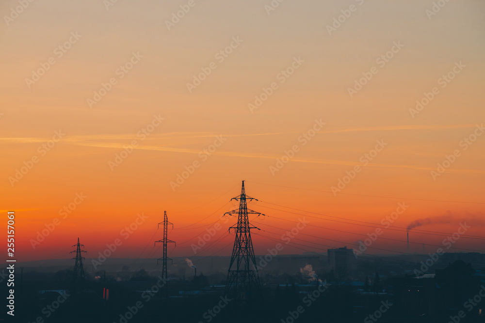 Power lines in city on dawn. Silhouettes of urban buildings among smog on sunrise. Cables of high voltage on warm orange yellow sky. Power industry at sunset. City power supply. Mist urban background.