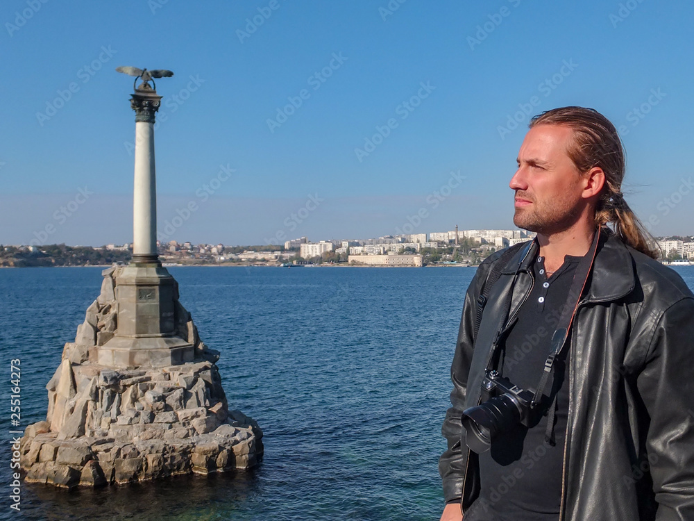 Young beautiful man photographer with blond long hair in black leather jacket with camera against Black Sea in right from Monument to Submerged Ships, the sybol of Sevastopol, Crimea.