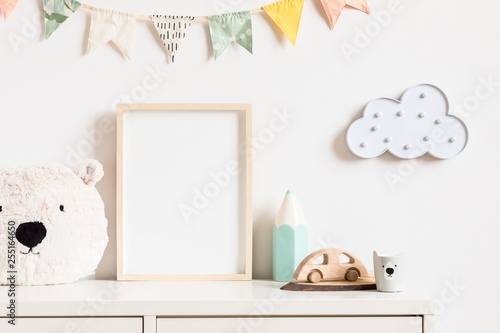 Stylish and modern scandinavian newborn baby interior with mock up photo or poster frame on the white shelf. Toys, teddy bear, wooden car and hanging cotton colorful flags and star. Template. Blank.