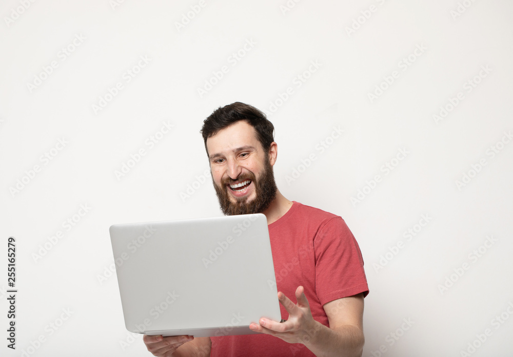 young bearded man looks at laptop screen in his hands and stands isolated on light background young bearded man looks at laptop screen in his hands and stands isolated on light background