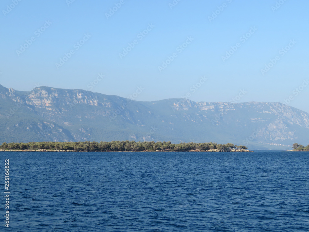 Sea coastline with mountain shore and rocky island covered with forest, view from the boat. Picturesque seascape with deep blue water in sunny day, beautiful panorama for background