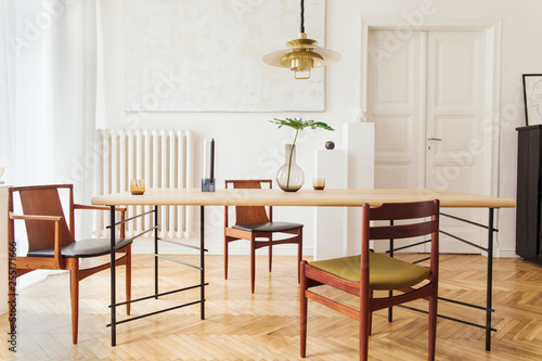 Eclectic and elegant dining room interior with design sharing table, chairs, gold pedant lamp, abstract paintings, piano and stylish accessories. Eclectic decor. Brown wooden parquet. Real photo.