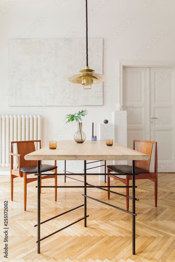 Fototapeta premium Eclectic and elegant dining room interior with design sharing table, chairs, gold pedant lamp, abstract paintings and stylish accessories. Tropical leafs in vase. Eclectic decor. Brown wooden parquet.