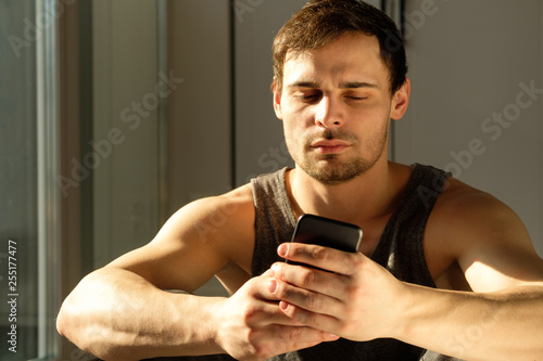 Young handsome man using phone while having exercise break in gym. Guy using smartphone after the daily training.