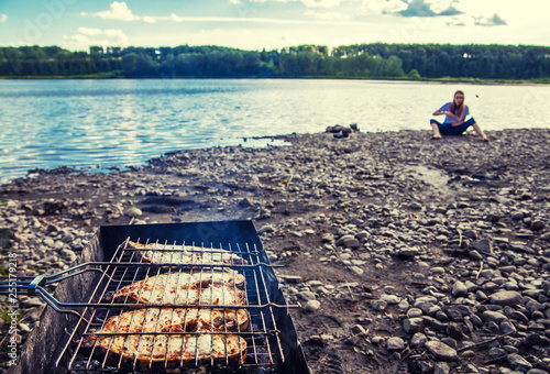 summer tourism by the river white clouds on the blue sky.  white fish steak is fried on the grill. in the background is a teenage girl in a white t-shirt sitting on the river Bank.