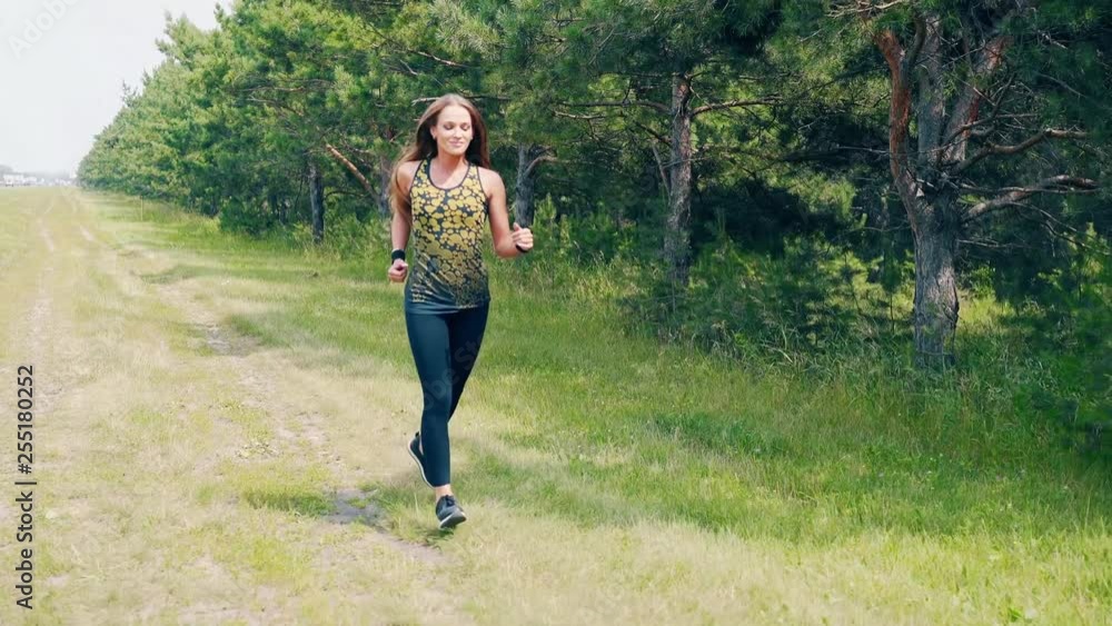 Young pretty girl goes in for sports in the morning, runs along pine forest. Wraps on her hands. She smiles. Young pretty girl goes in for sports in the morning, runs along pine forest. Wraps on her hands. She smiles.