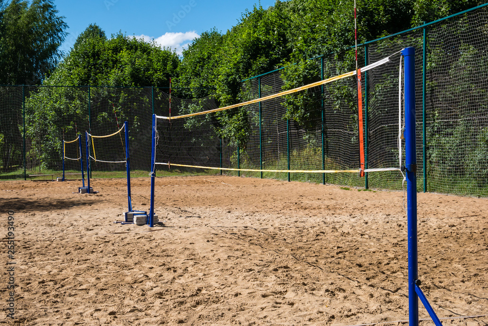 Rows of volleyball nets on blue poles on empty sand court surrounded by lush green trees on summer day