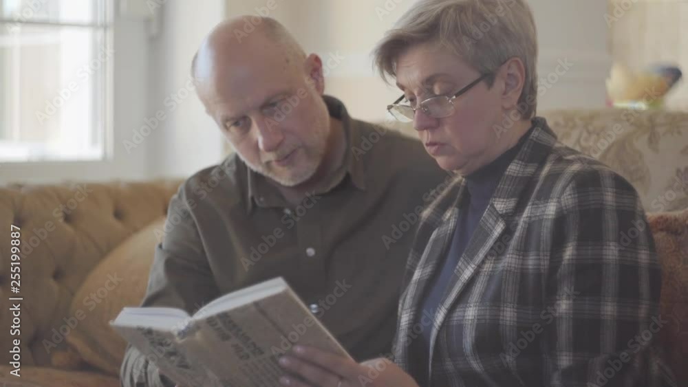 Elder couple, man and woman, sitting on the sofa with a book and talking and smiling to each other. Family relationships. Elder couple, man and woman, sitting on the sofa with a book and talking and smiling to each other. Family relationships.