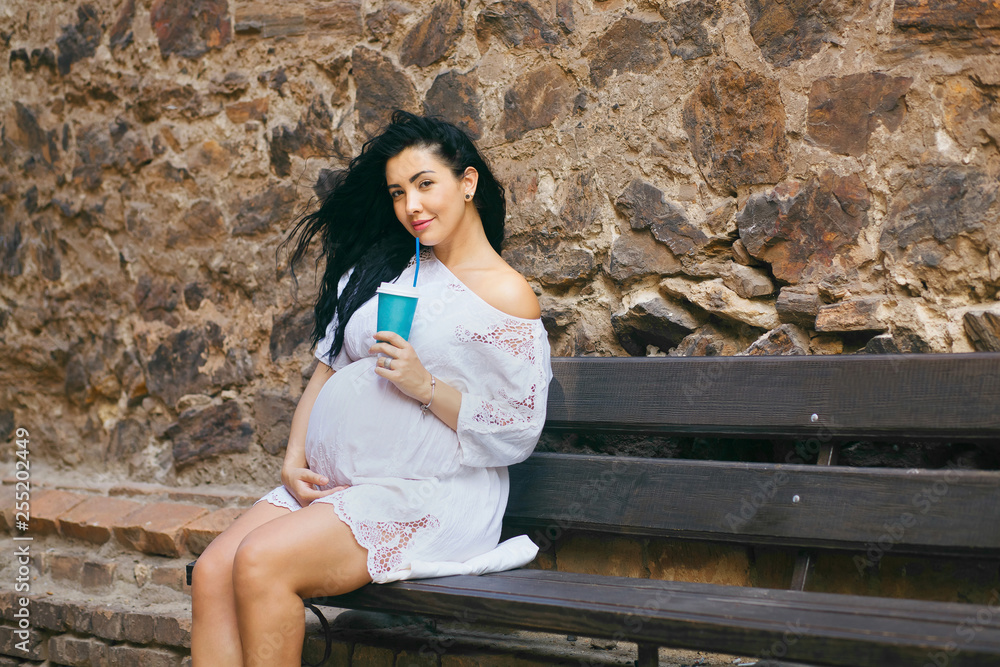 a pregnant girl sits in a romantic place, smiles and drinks warm tea, a beautiful girl sits on a bench against the background of a stone wall