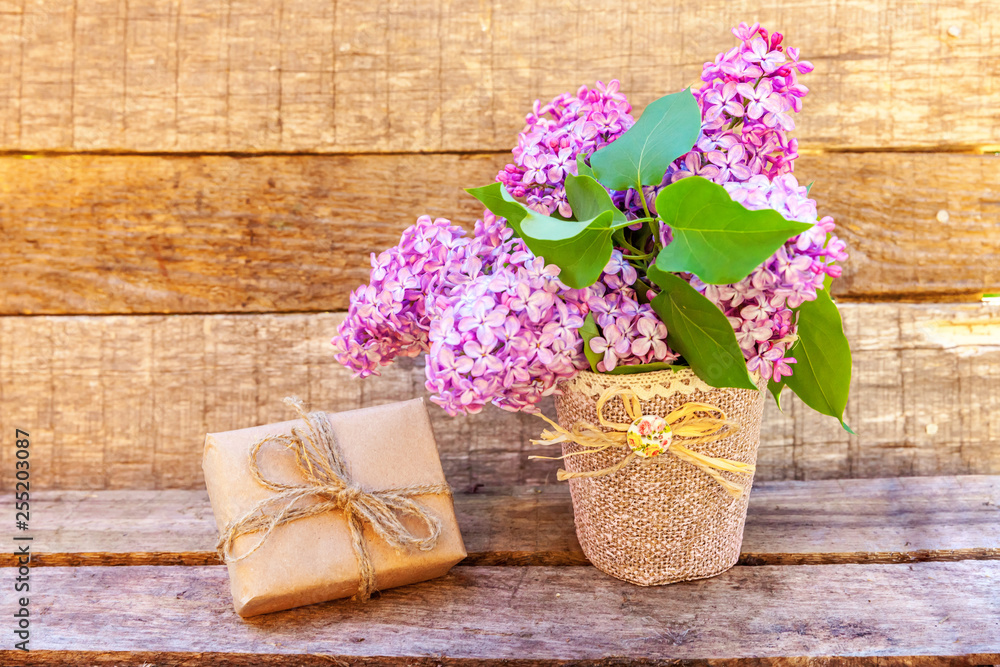 Floral decor elements. Small gift box with presents wrapped craft brown paper with bouquet of flowers beautiful smell violet purple lilac in vase on rustic wooden background. Copy space Floral decor elements. Small gift box with presents wrapped craft brown paper with bouquet of flowers beautiful smell violet purple lilac in vase on rustic wooden background. Copy space