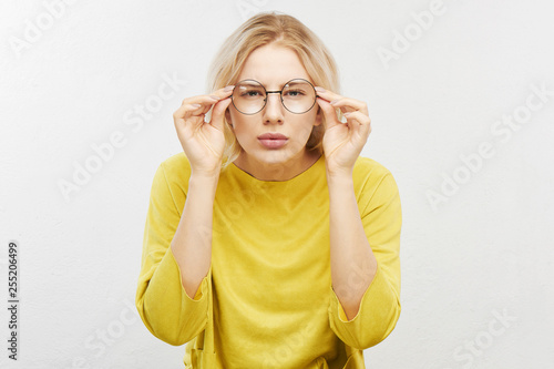 Portrait of a beautiful young woman with spectacles, looking curiously at you, squinting, trying to look closer. Girl with poor eyesight in yellow T-shirt posing on a white background in the studio