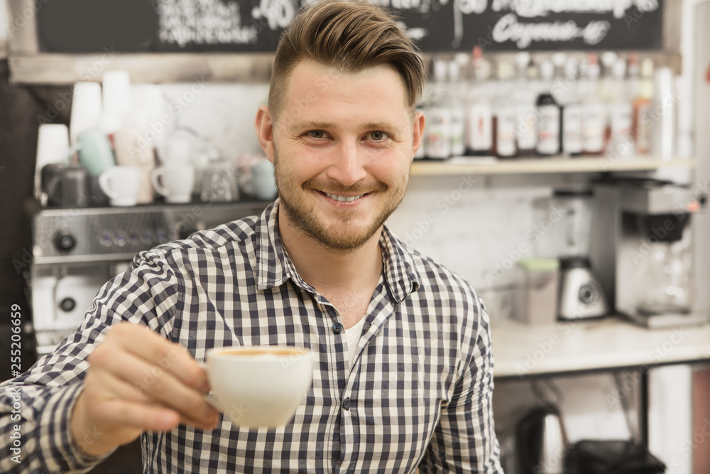 Handsome young male barista smiling cheerfully holding out a cup of fresh delicious coffee to the camera working at his coffee shop copyspace small business owner offering beverage positivity friendly