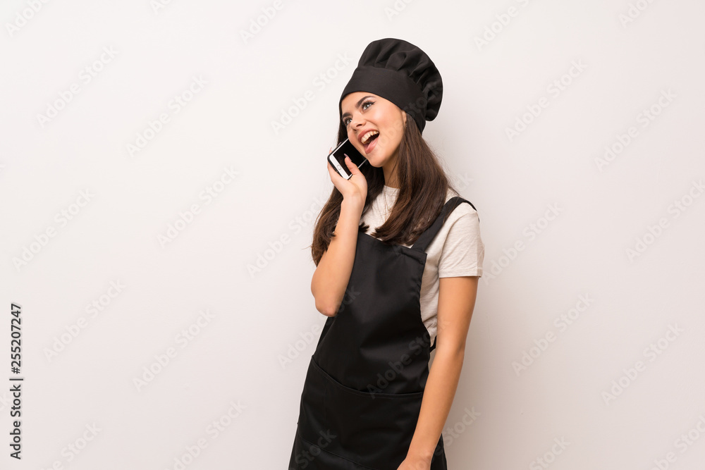 Teenager chef over white wall keeping a conversation with the mobile phone Teenager chef over white wall keeping a conversation with the mobile phone