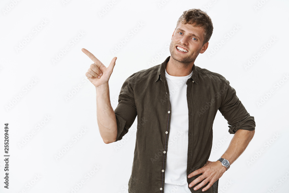 Pleasant and charismatic blond boyfriend pointing left as showing around new apratment smiling friendly at camera holding hand on waist during casual relaxing conversation over gray background Pleasant and charismatic blond boyfriend pointing left as showing around new apratment smiling friendly at camera holding hand on waist during casual relaxing conversation over gray background