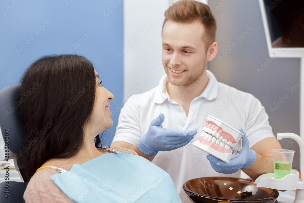 Obraz premium Teeth care. Handsome young professional dentist explaining his senior patient how to care after teeth showing a teeth mold working at dental clinic professional communication medical service concept