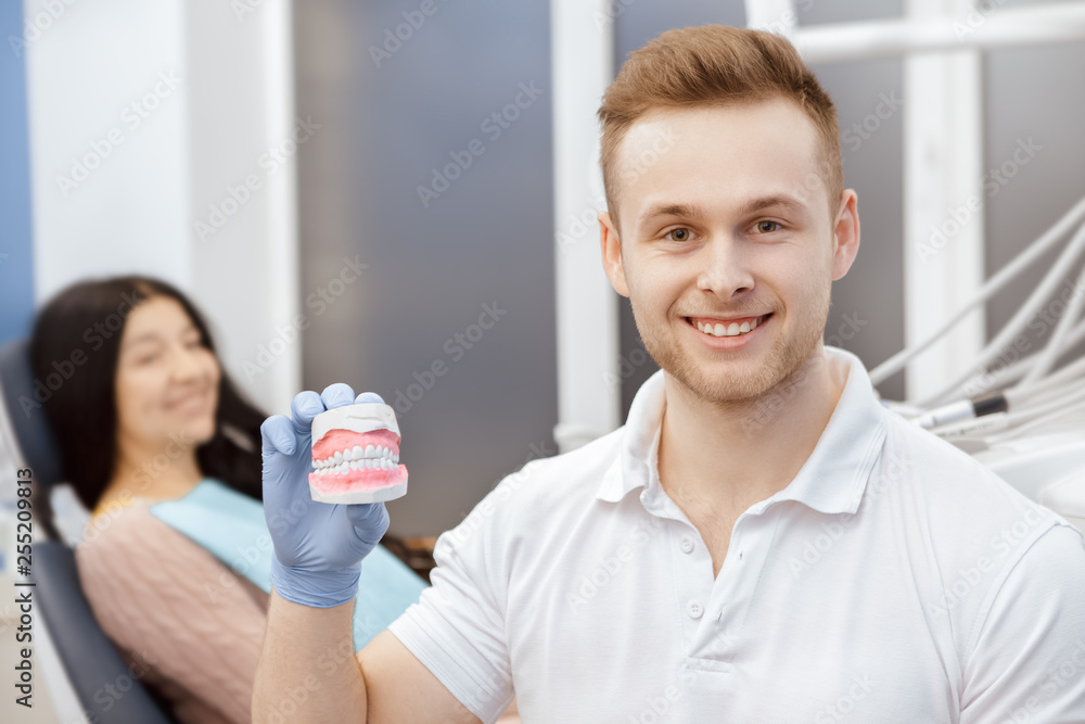 He cares for your health. Cheerful professional dentist smiling to the camera holding dental mold his patient sitting on the back copyspace healthcare medicine professionalism perfect smile concept He cares for your health. Cheerful professional dentist smiling to the camera holding dental mold his patient sitting on the back copyspace healthcare medicine professionalism perfect smile concept