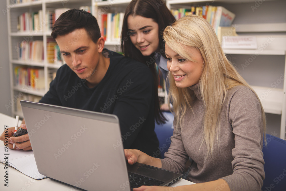 Happy beautiful female student smiling, talking to her college friends, while working on a project togther at the campus library. Group of high school friends studying, using computer Happy beautiful female student smiling, talking to her college friends, while working on a project togther at the campus library. Group of high school friends studying, using computer