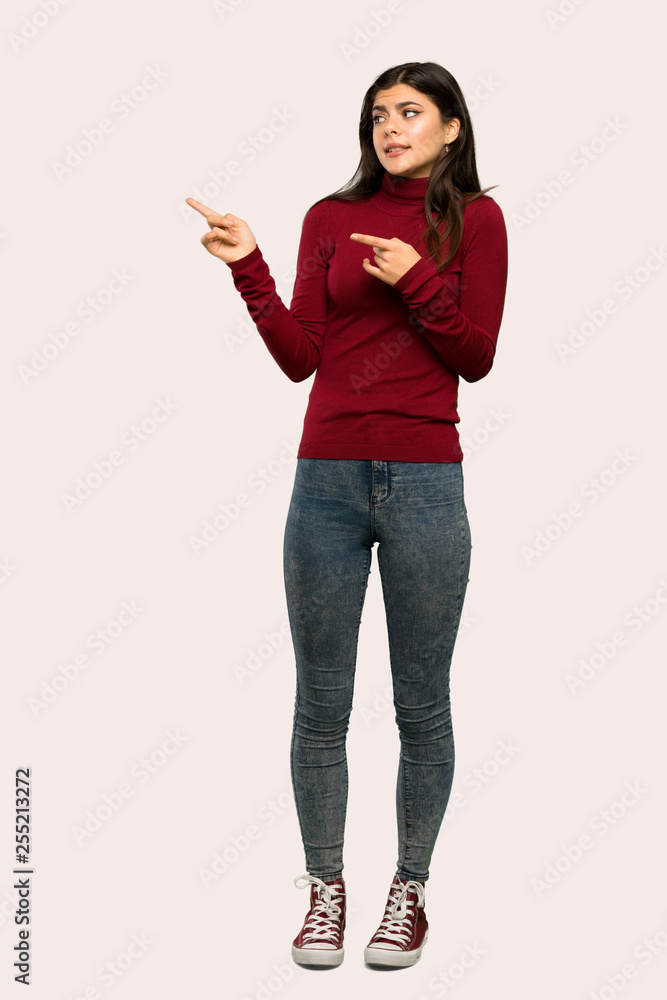 A full-length shot of a Teenager girl with turtleneck frightened and pointing to the side over isolated background A full-length shot of a Teenager girl with turtleneck frightened and pointing to the side over isolated background