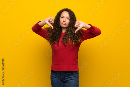 Teenager girl with red sweater over yellow wall showing thumb down