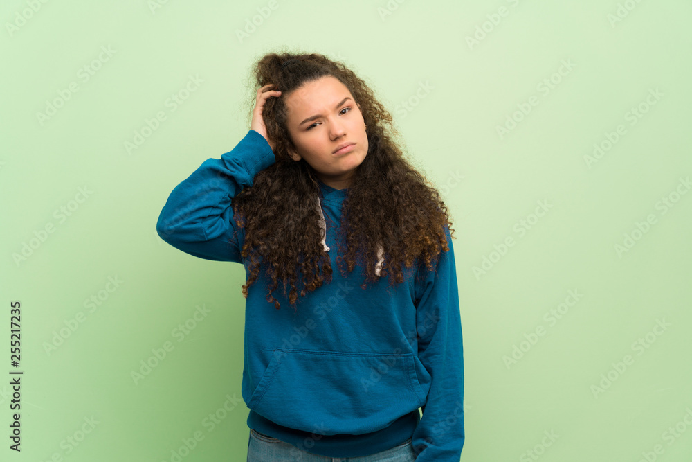 Teenager girl over green wall having doubts while scratching head Teenager girl over green wall having doubts while scratching head