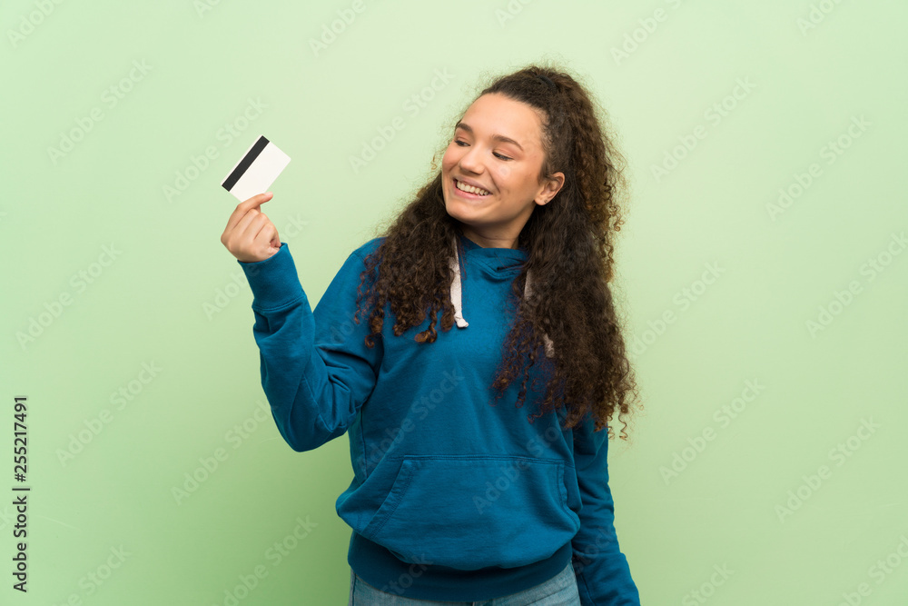 Teenager girl over green wall holding a credit card and thinking Teenager girl over green wall holding a credit card and thinking