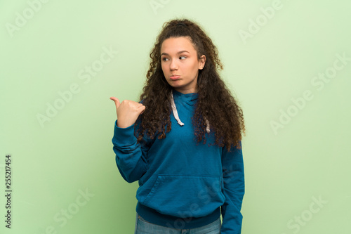 Teenager girl over green wall unhappy and pointing to the side © luismolinero