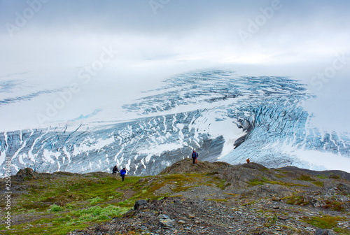 End of the trail. Tourists looking at the view. Closed clouds day, overlooking the Exit Glacier showing its blue ice, white snow, and a lush landscape. In Kenai Fjords National Park - July 27, 2018