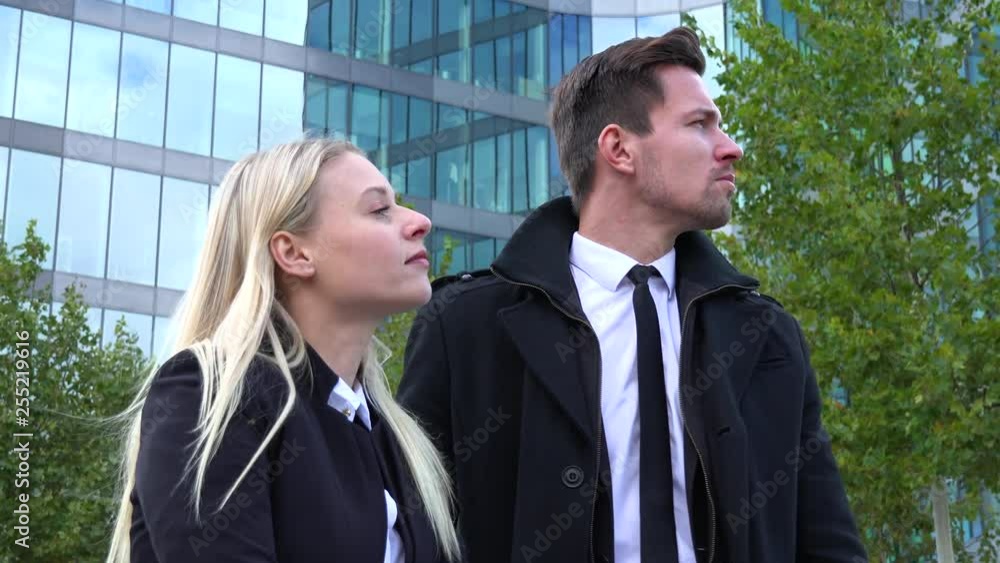 A businessman and a businesswoman stand in front of an office building and look around - view from below