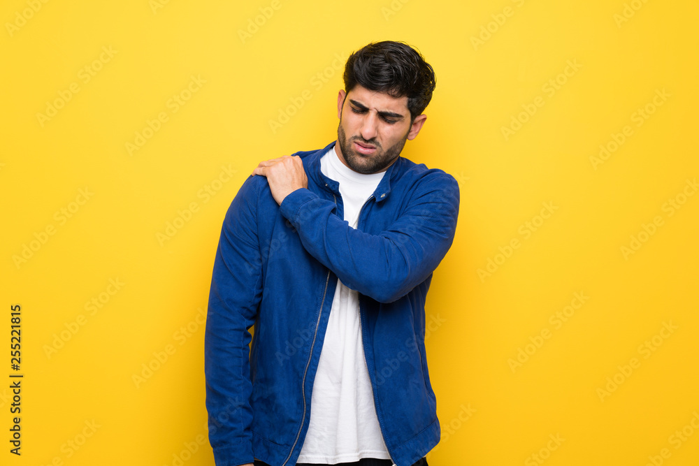 Man with blue jacket over yellow wall suffering from pain in shoulder for having made an effort Man with blue jacket over yellow wall suffering from pain in shoulder for having made an effort