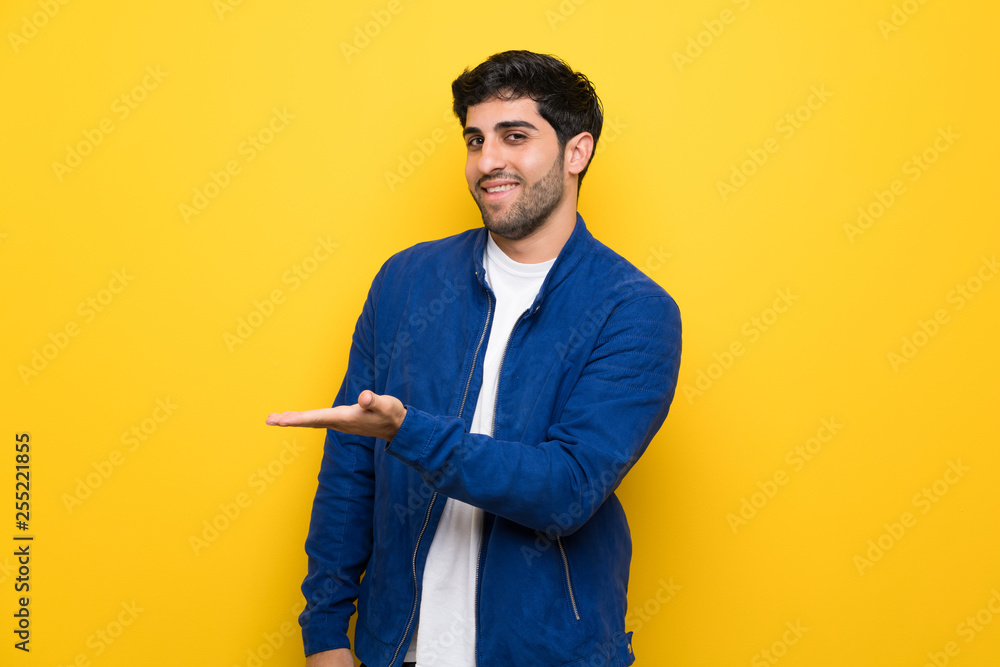 Man with blue jacket over yellow wall presenting an idea while looking smiling towards