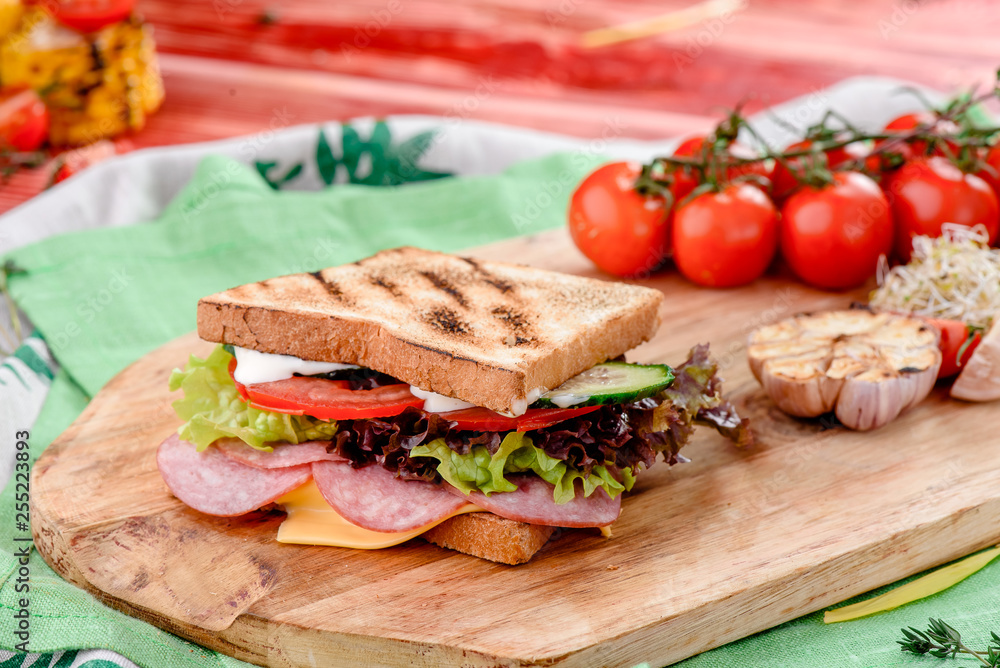 sandwich with sausage, tomatoes, onions, lettuce and cheese on a wooden board on a red wooden background, decorated with napkins, chili pepper and cherry tomatoes. close-up. close up. space