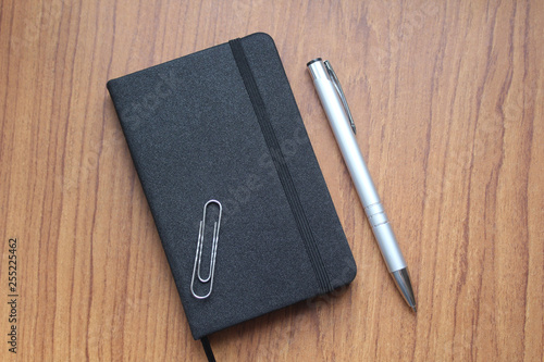 Black notebook for records, an office paper clip and a ball pen of silver color on a table. Business subject. It is used in a web design.