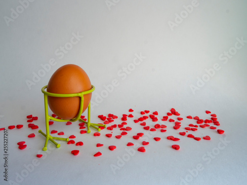 Brown egg of light color in a support prepares for the holiday Easter on a white background strewed with bright red hearts.