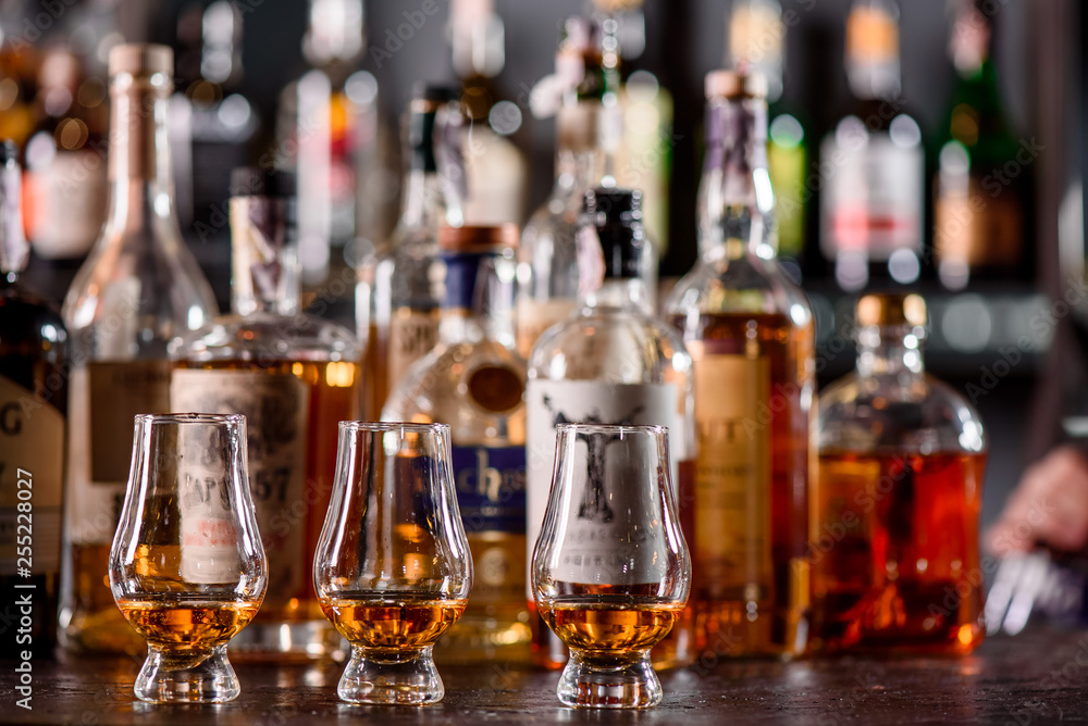 Three Glencairn Glass with whiskey on a bar wooden counter close up on the background of blurry bottles. Close up. Dark background Three Glencairn Glass with whiskey on a bar wooden counter close up on the background of blurry bottles. Close up. Dark background