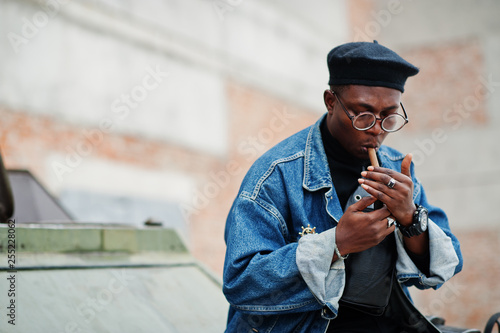 African american man in jeans jacket, beret and eyeglasses, lights a cigar and posed against btr military armored vehicle.