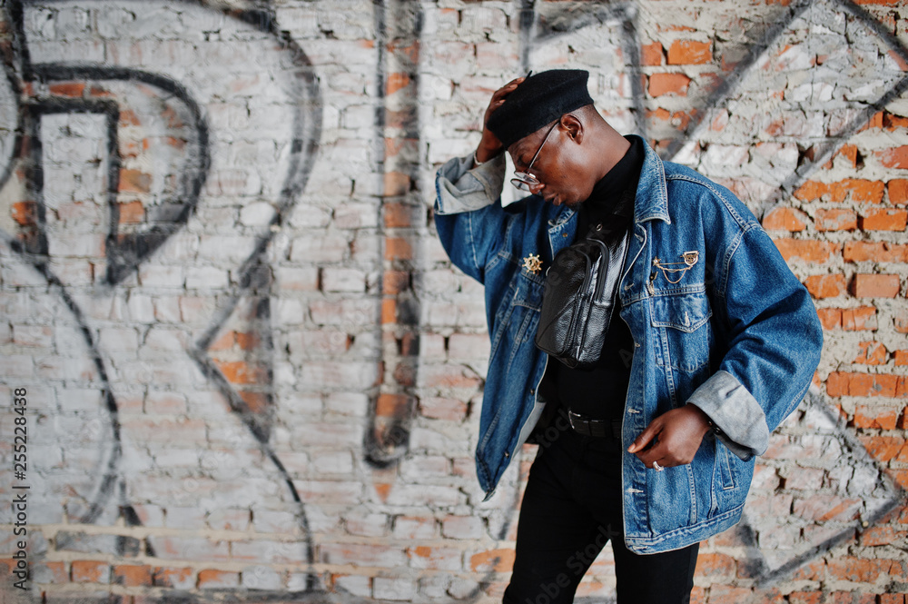 African american man in jeans jacket, beret and eyeglasses against graffiti brick wall with bruk sign. - obrazy, fototapety, plakaty 