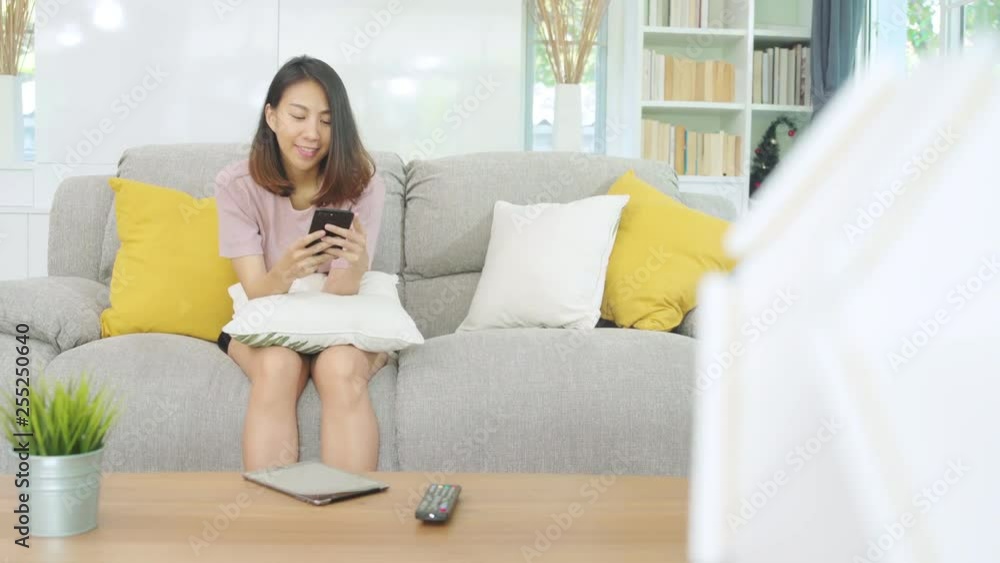 Young Asian woman using smartphone checking social media feeling happy smiling while lying on the sofa when relax in living room at home. Lifestyle latin and hispanic ethnicity women at house concept. Young Asian woman using smartphone checking social media feeling happy smiling while lying on the sofa when relax in living room at home. Lifestyle latin and hispanic ethnicity women at house concept.