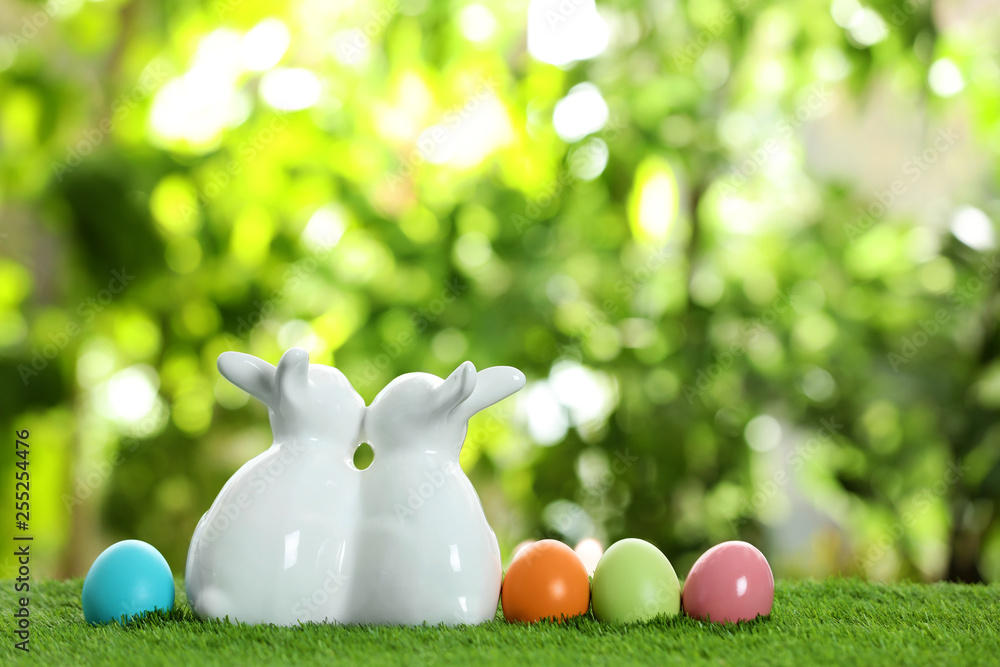 Cute ceramic Easter bunnies and dyed eggs on green grass against blurred background, space for text Cute ceramic Easter bunnies and dyed eggs on green grass against blurred background, space for text