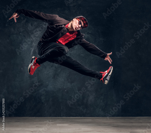 Emotional stylish dressed guy performing break dance jumping. Studio photo against a dark textured wall