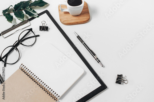 Home office workspace mockup with blank clip board, office supplies, pen, green leaf, coffee cup on a wooden stand and eyeglasses on white background. Flat lay, top view, copy space