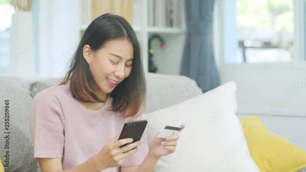 Young smiling Asian woman using smartphone buying online shopping by credit card while lying on sofa when relax in living room at home. Lifestyle latin and hispanic ethnicity women at house concept. Young smiling Asian woman using smartphone buying online shopping by credit card while lying on sofa when relax in living room at home. Lifestyle latin and hispanic ethnicity women at house concept.